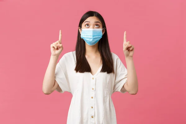 Covid-19, socialt distanserande, virus och livsstil. Nyfiken söt asiatisk flicka i medicinsk mask och sommarklänning, tittar och pekar finger upp på promo, reklam, rosa bakgrund — Stockfoto