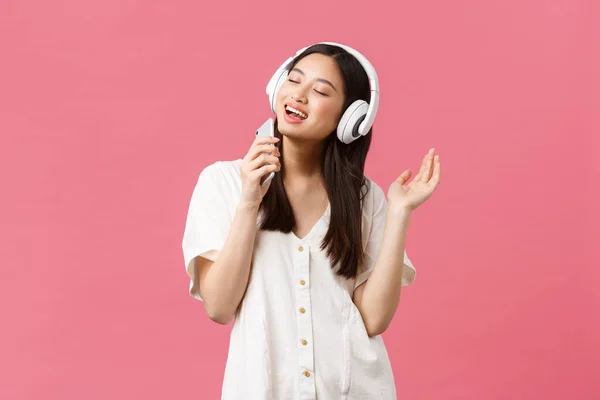 Belleza, emociones de la gente y concepto de tecnología. Despreocupado chica asiática feliz usando aplicación de karaoke de teléfono móvil, cantando en el micrófono del teléfono inteligente, escuchar música en los auriculares, fondo rosa — Foto de Stock