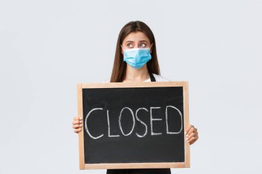 Covid-19 social distancing, cafe employees, coffee shops and coronavirus concept. Upset cute female barista in medical mask looking away sad as showing we are closed sign, cant open during pandemic clipart
