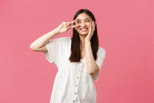 Beleza, emoções das pessoas e conceito de lazer de verão. Sonhador e romântico bonito sorrindo menina asiática olhando afastado pensativo ao tocar pele macia no rosto e mostrar sinal de paz, fundo rosa — Fotografia de Stock