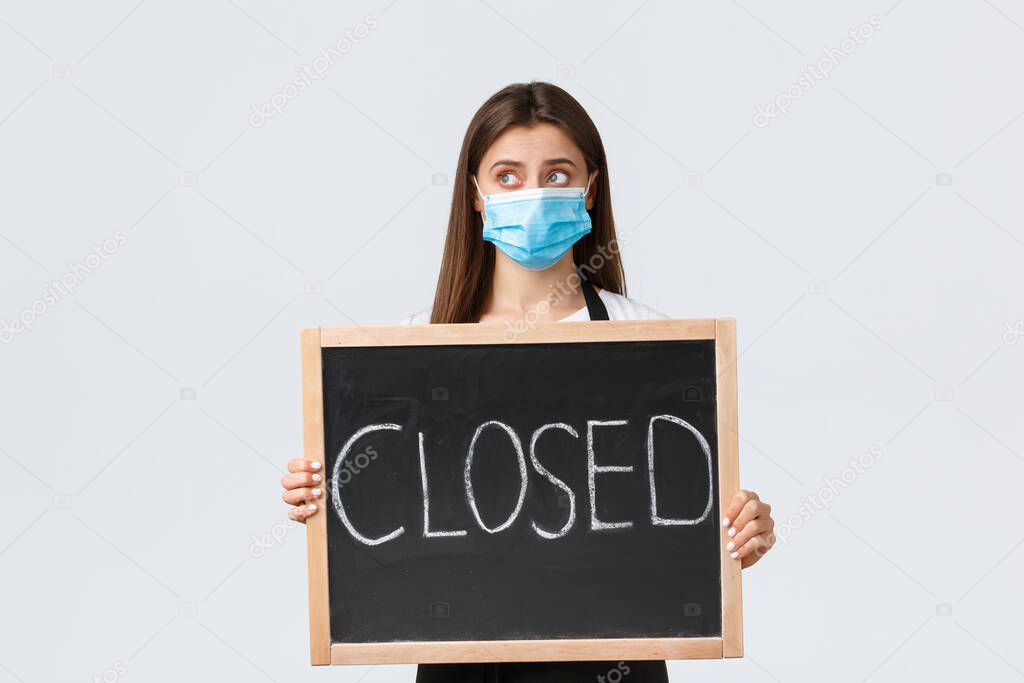 Covid-19 social distancing, cafe employees, coffee shops and coronavirus concept. Upset cute female barista in medical mask looking away sad as showing we are closed sign, cant open during pandemic