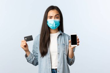 Sosyal mesafeli yaşam tarzı, covid-19 salgını ve temassız alışveriş konsepti. Şüpheli ve şüpheli maskeli bir kadın cep telefonu ekranı ve kredi kartını şüpheli bir ifadeyle gösteriyor.