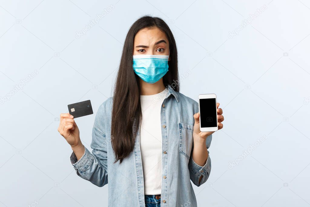 Social distancing lifestyle, covid-19 pandemic and contactless shopping concept. Skeptical and suspicious woman in medical mask showing mobile phone screen and credit card with doubtful expression