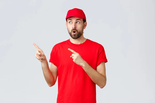 Online shopping, delivery during quarantine and takeaway concept. Impressed and wondered courier in red t-shirt with cap, employee uniform, open mouth shocked and staring left, pointing fingers — Stock Photo, Image