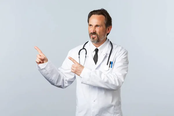 Covid-19, coronavirus outbreak, healthcare workers and pandemic concept. Mad annoyed doctor cursing, pointing fingers upper left corner and frowning disapproval, complaining — Stock Photo, Image