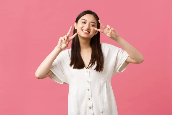 Schönheit, Menschen-Emotionen und Sommerfreizeitkonzept. Dumm und zart lächelnd, glückliches japanisches Mädchen in weißem Kleid, mit Peace-Kawaii-Zeichen und grinsendem fröhlichem, stehendem rosa Hintergrund — Stockfoto