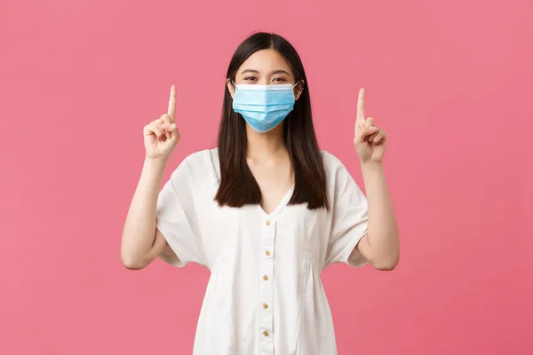 Covid-19, socialt distanserande, virus och livsstil. Glada leende kvinna i snygg vit klänning, sommar outfit och medicinsk mask, peka finger upp, främja reklam, rosa bakgrund — Stockfoto