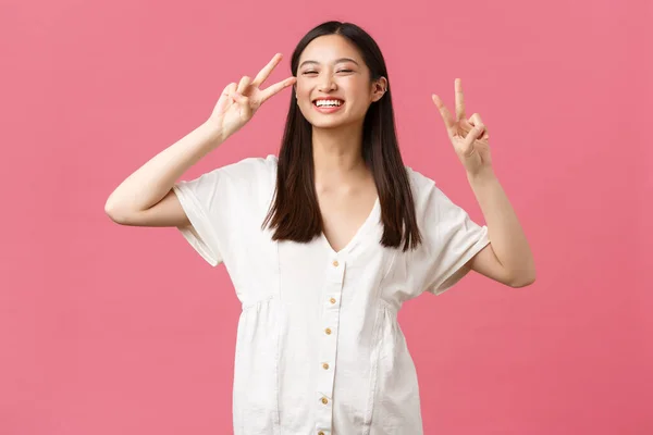 Belleza, emociones de la gente y concepto de ocio de verano. alegre kawaii asiático chica en blanco lindo vestido, mostrando paz signgs y sonriendo, de pie optimista sobre rosa fondo —  Fotos de Stock