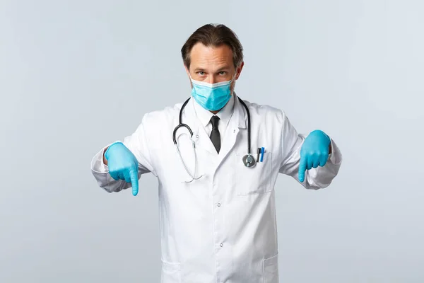 Covid-19, πρόληψη του ιού, εργαζόμενοι στον τομέα της υγείας και την έννοια του εμβολιασμού. Σοβαρός γιατρός με ιατρική μάσκα και γάντια, δείχνοντας τα δάχτυλα προς τα κάτω, δείχνοντας promo banner, λευκό φόντο — Φωτογραφία Αρχείου