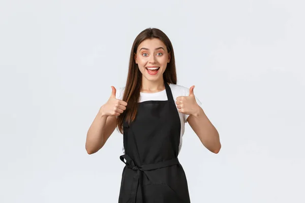 Grocery store employees, small business and coffee shops concept. Excited cheerful female barista show thumbs-up in approval. Saleswoman in black apron recommending product or special drink