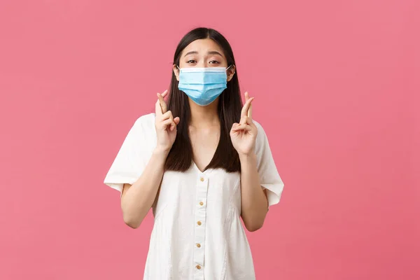 Covid-19, socialt distanserande, virus och livsstil. Förhoppningsfull drömsk asiatisk flicka att önska, förutse nyheter i medicinsk mask, kors fingrar lycka till, vädjar över rosa bakgrund — Stockfoto