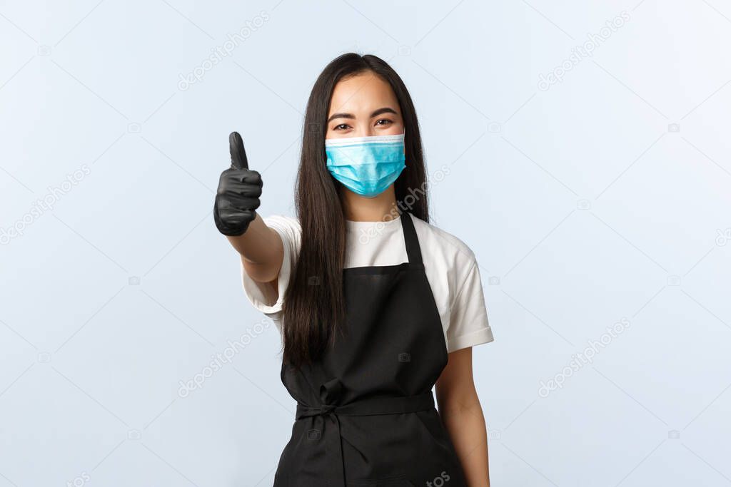 Covid-19, social distancing, small coffee shop business and preventing virus concept. Smiling asian female barista, employee in medical mask and gloves show thumb-up