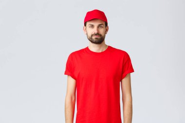 İnternetten alışveriş, karantina sırasında teslimat ve paket servis konsepti. Kırmızı üniformalı, şapkalı ve tişörtlü genç bir kurye kameraya bakıyor, siparişinizi bekliyor.