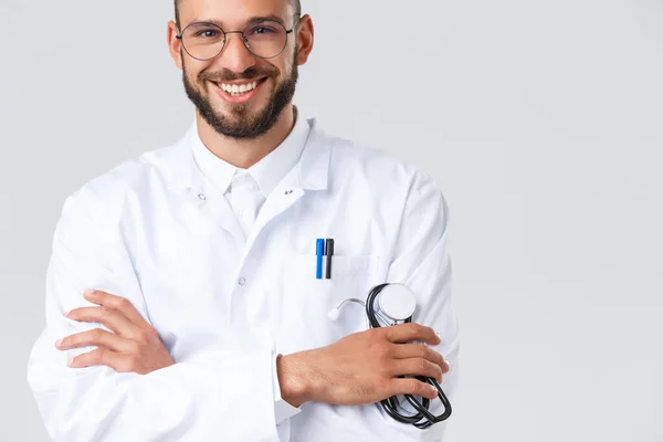 Healthcare workers, coronavirus, covid-19 pandemic and insurance concept. Close-up of handsome smiling male doctor in white coat, glasses, cross arms chest and hold stethoscope, talk to patients — Stock Photo, Image