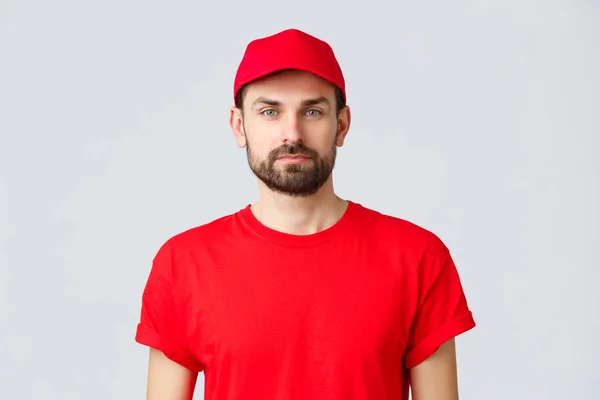 Nätshopping, leverans under karantän- och hämtningskoncept. Ung snygg skäggig kurir i röd serviceuniform med mössa och t-shirt, anställd som arbetar överföra order — Stockfoto