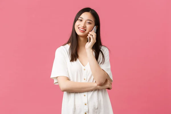 Belleza, emociones de la gente y concepto de tecnología. Feliz chica asiática en vestido blanco hablando por teléfono, la celebración de móvil y la mirada de la cámara, llamando a un amigo, fondo rosa — Foto de Stock