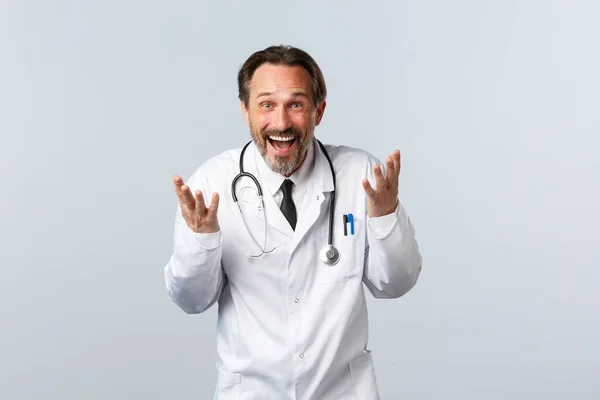 Covid-19, brote de coronavirus, trabajadores sanitarios y concepto pandémico. Emocionado médico varón feliz riendo y explicando algo divertido, sonriendo ampliamente, de pie fondo blanco — Foto de Stock