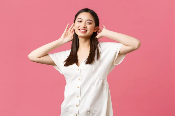 Belleza, emociones de la gente y concepto de ocio de verano. Relajada y feliz chica asiática en vestido, disfrutando de un día soleado brillante de vacaciones, cogidas de la mano cerca de la cara y sonriente, fondo rosa — Foto de Stock