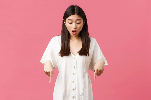 Skönhet, människor känslor och sommar fritid och semester koncept. Överraskad och upphetsad kawaii asiatisk flicka i vit klänning, peka och titta ner med roade glad ansikte, rosa bakgrund — Stockfoto