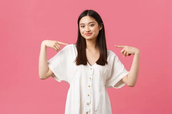 Belleza, emociones de la gente y el ocio de verano y el concepto de vacaciones. Confiada chica asiática con estilo en vestido blanco, sonriendo satisfecha y apuntando a sí misma orgullosa, presumida, de pie fondo rosa — Foto de Stock