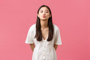 Güzellik, insan duyguları ve yaz tatili konsepti. Beyaz romantik elbiseli sevimli Asyalı kız, öpücük vermek için öne eğiliyor, pembe arka planda bekliyor.