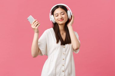 Güzellik, insan duyguları ve teknoloji konsepti. Tasasız ve güzel Asyalı kız kablosuz kulaklıklarla müzik dinliyor, gözleri kapalı gülümsüyor en sevdiği şarkıyı dinliyor, akıllı telefon tutuyor ve dans ediyor.