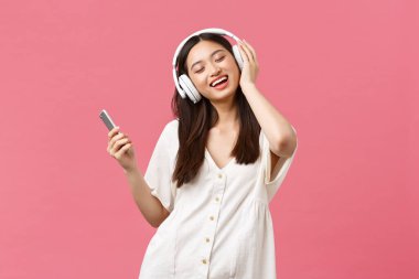 Güzellik, insan duyguları ve teknoloji konsepti. Tasasız ve güzel Asyalı kız kablosuz kulaklıklarla müzik dinliyor, gözleri kapalı gülümsüyor en sevdiği şarkıyı dinliyor, akıllı telefon tutuyor ve dans ediyor.