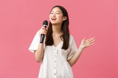 Boş zaman, insanların duyguları ve yaşam tarzı konsepti. Tasasız güzel Asyalı kız şık giyinmiş, mikrofonda şarkı söylüyor, karaoke gecesinin tadını çıkarıyor, pembe arka planda eğleniyor.