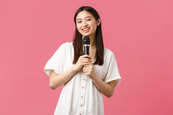 Lazer, emoções das pessoas e conceito de estilo de vida. alegre sorrindo asiático menina no karaoke, aproveitando fins de semana, canto canção no microfone, executar stand-up, pé rosa fundo — Fotografia de Stock