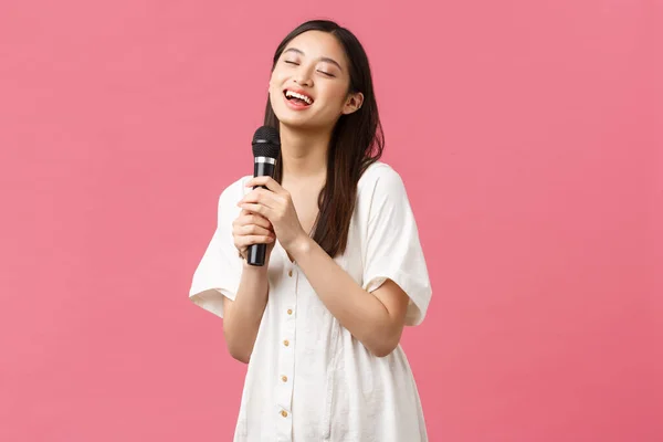 Lazer, emoções das pessoas e conceito de estilo de vida. Carefree menina asiática bonita em vestido elegante, cantando música no microfone, desfrutando de noite de karaoke, se divertindo sobre fundo rosa — Fotografia de Stock