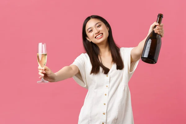 Celebration, party holidays and fun concept. Happy smiling asian girl host of event, holding bottle champagne and glass and reaching hands for hugging guest, standing pink background