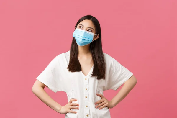 Covid-19, socialt distanserande, virus och livsstil. Drömmande söt asiatisk flicka i vit klänning och medicinsk mask mot coronavirus, tittar övre vänstra hörnet avbildning, ler glad — Stockfoto