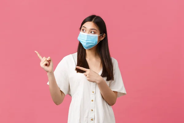 Covid-19 、社会的距離、ウイルスやライフスタイルの概念。好奇心旺盛なアジア系の女の子,医療用マスクに訪問者を格納製品についての質問,指差し指を左,立ってピンクの背景 — ストック写真
