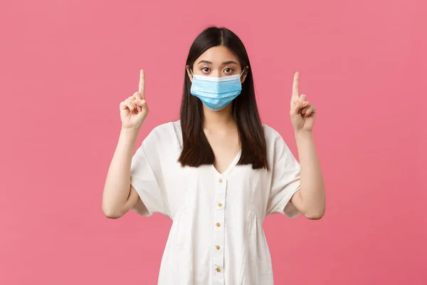 Covid-19, social distancing, virus and lifestyle concept. Pretty stylish asian girl in medical mask suggest visit this store or place, pointing fingers up at product, standing pink background — Stock Photo, Image
