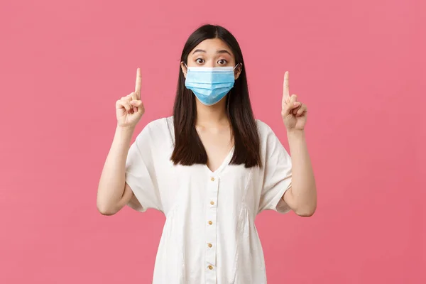 Covid-19, socialt distanserande, virus och livsstil. Förvånad asiatisk flicka i medicinsk mask ser kameran fascinerad och ställa frågan om produkten på toppen, peka finger upp, rosa bakgrund — Stockfoto