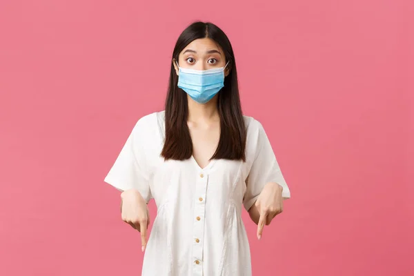 Covid-19, socialt distanserande, virus och livsstil. Spännande och fascinerad söt kvinnlig butik kund i medicinsk mask, ställa frågan om produkt promo erbjudande, peka finger ner — Stockfoto