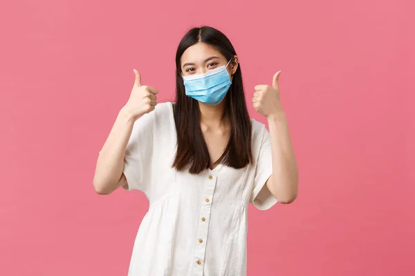 Covid-19, social distancing, virus and lifestyle concept. Upbeat pleased attractive asian woman in white dress and medical mask staying safe during coronavirus, showing thumbs-up — Stock Photo, Image