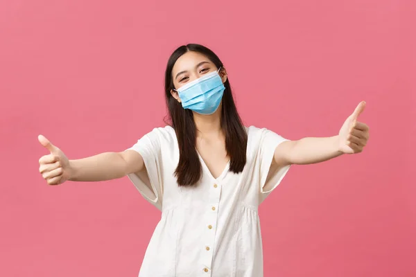 Covid-19, társadalmi távolság, vírus és életmód koncepció. Barátságos boldog ázsiai nő orvosi maszk és nyári ruha, kinyújtja a kezét, hogy ölelést, üdvözöl valakit, álló rózsaszín háttér — Stock Fotó