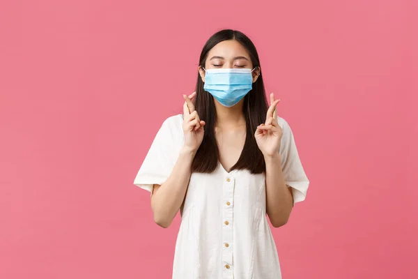 Covid-19, socialt distanserande, virus och livsstil. Hopplös drömsk asiatisk flicka som gör önskan, blunda och ler i medicinsk mask, kors fingrar lycka till, vädjar över rosa bakgrund — Stockfoto