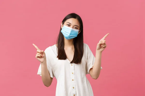 Covid-19, soziale Distanzierung, Virus- und Lebensstilkonzept. Charismatische Kawaii-Japanerin in medizinischer Maske und Sommerkleid zeigt mit dem Finger zur Seite und zeigt links und rechts Sonderangebote — Stockfoto