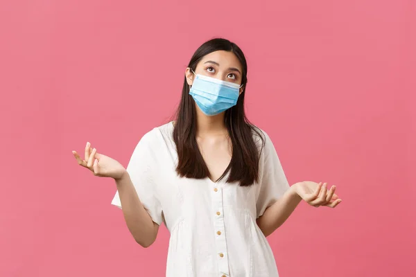 Covid-19, sociale afstand, virus en lifestyle concept. Onnozele en aarzelende jonge aziatische vrouw met medisch masker, shrugging en opkijken twijfelachtig, wegen keuzes, staande roze achtergrond — Stockfoto