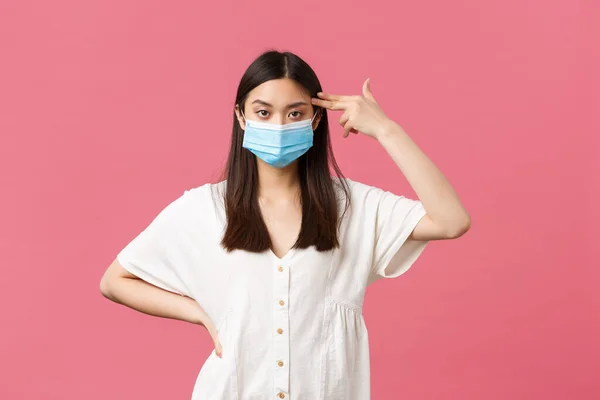 Covid-19, social distancing, virus and lifestyle concept. Annoyed and distressed asian female student in medical mask, showing fake gun over temple as shooting herself from boredom or annoyance