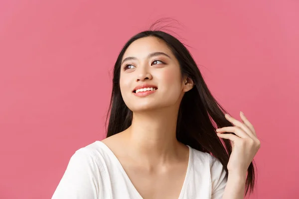 Publicidade de produtos de beleza, cuidados com o cabelo e conceito de moda feminina. Romântico concurso mulher asiática com belo sorriso branco, olhando longe sonhador como tocar o cabelo, de pé fundo rosa — Fotografia de Stock