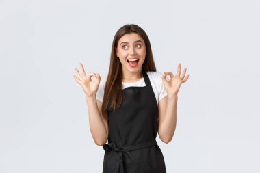 Market çalışanları, küçük işletmeler ve kahve dükkanları konsepti. Heyecanlı, siyah önlüklü, rüya gibi kadın barista şaşırmış görünüyor ve harika bir içki önermek için iyi bir jest yapıyor.