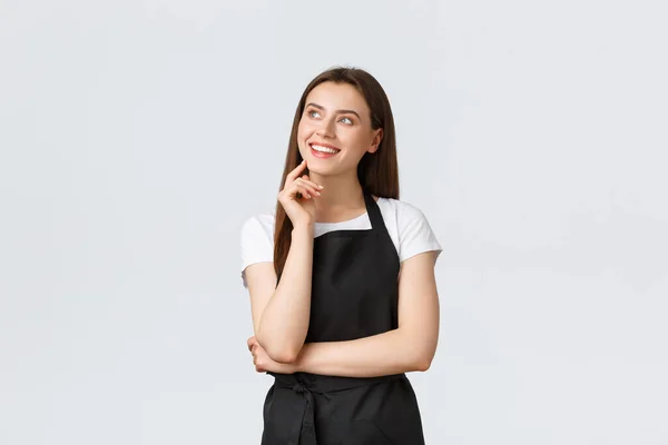 Kleinunternehmen, Mitarbeiter und Café-Konzept. Verträumt lächelnde schöne Verkäuferin in schwarzer Schürze, die oben rechts aussieht. Verkäuferin oder Kellner im Restaurant — Stockfoto