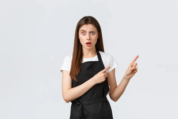 Small business, employees and coffee shop concept. Confused barista in black apron frowning and looking concerned as pointing upper right corner worried, standing white background — Stock Photo, Image