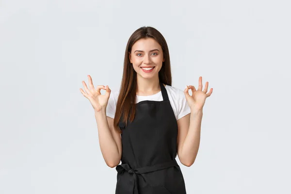 Les employés des épiceries, les petites entreprises et les cafés concept. Souriant vendeuse amicale en tablier noir montrant un geste correct, recommander ou garantir la qualité des boissons — Photo