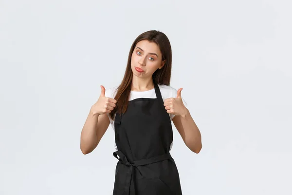 Grocery store employees, small business and coffee shops concept. Excited cheerful female barista show thumbs-up in approval. Saleswoman in black apron impressed with cool new promo
