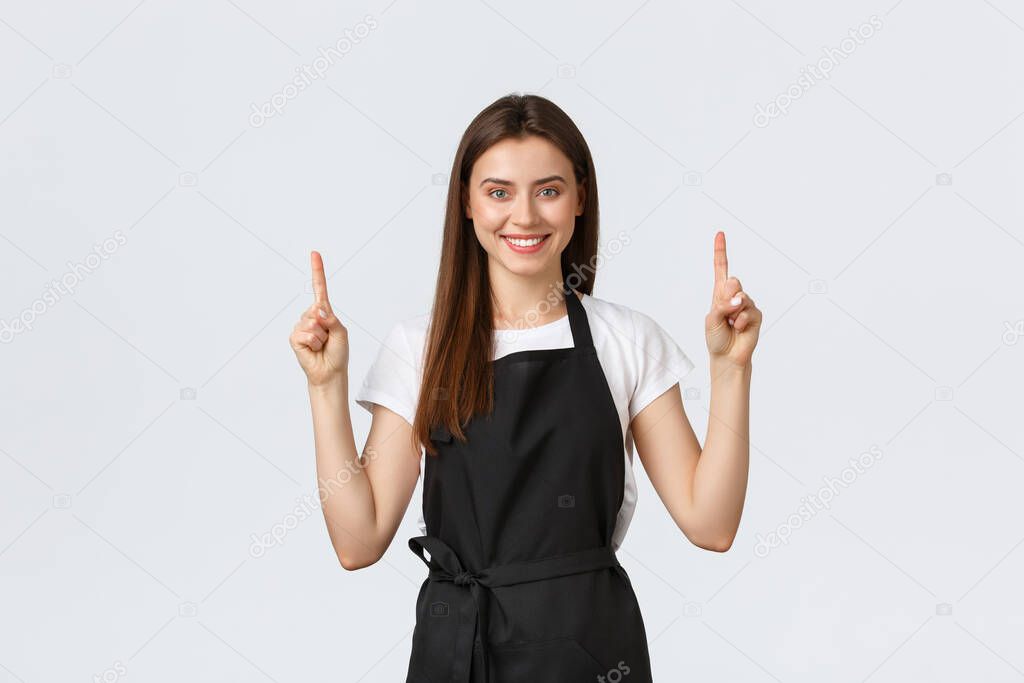 Small business, employees and coffee shop concept. Confient pretty barista in black apron pointing fingers up. Cafe employee showing advertisement. Cheerful saleswoman recommend special promo