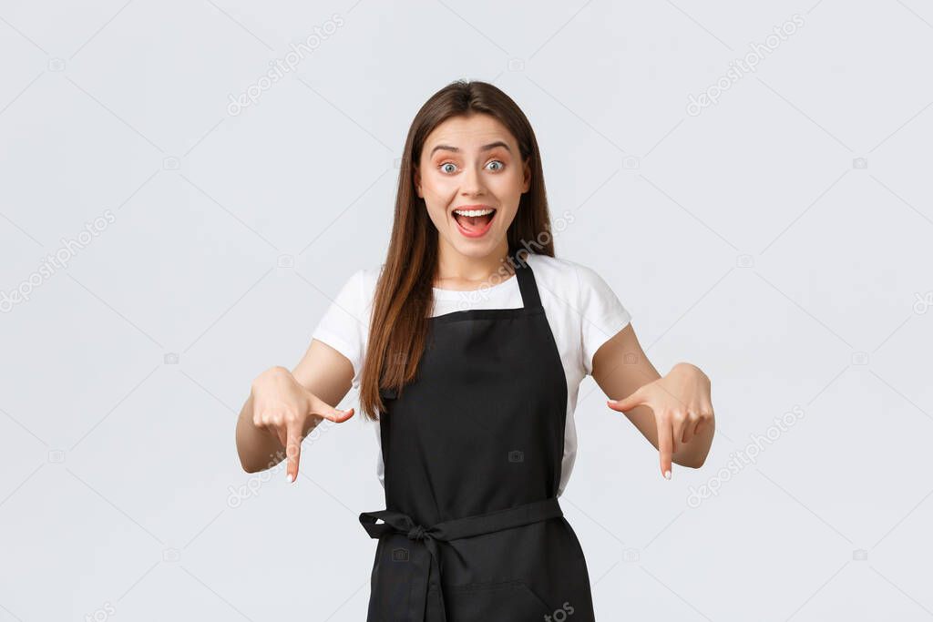 Small business, employees and coffee shop concept. Excited and impressed grocery store saleswoman in black apron pointing fingers down astonished. Cute cafe worker provide information